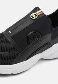 Sorte sneaker med mesh og glatt syntetisk overdel, en frontglidelås med gullakenter og en hvit teksturert såle. Sirkulært logodetalj.