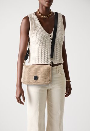 Borsa a tracolla intrecciata beige con un logo circolare nero, caratterizzata da una superficie testurizzata e una tracolla nera regolabile. Abbinata a un abbigliamento di colore crema.