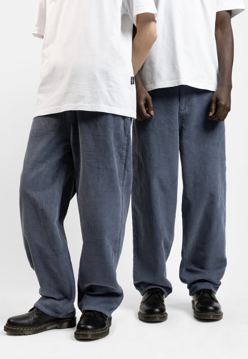 Pantaloni a gamba larga in corduroy blu con una trama a coste, abbinati a scarpe nere con cuciture gialle. Due modelli sono in piedi uno accanto all'altro.