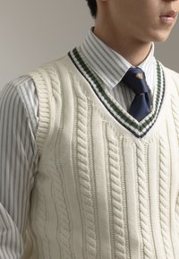 Chaleco de punto grueso color crema con escote en V, sobre una camisa rayada. El chaleco cuenta con acentos en verde y marino alrededor del escote.