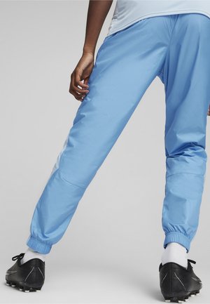 Pantalones deportivos de color azul claro con puños elásticos, franjas laterales blancas y un corte relajado. Combinados con botas de fútbol negras y calcetines blancos.
