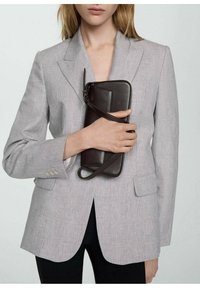 Lysgrå plaid-blazer med spidse klapper og to frontlommer, holdt med den ene hånd over en brun læderclutch med en rem.