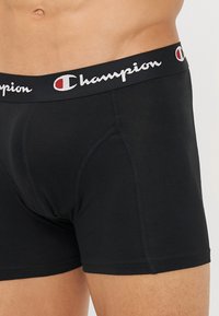 Black cotton boxer briefs with a thick elastic waistband featuring the Champion logo in white and red. Flatlock seams provide a smooth fit.