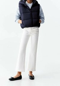 Navy puffer vest with hood, light blue knitted sweater, white flared jeans, and black flat shoes with bow detail.