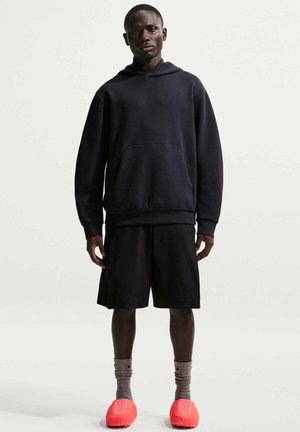 Ung mand iført en sort hættetrøje, sorte shorts, grå sokker og klare røde slip-on sko, der står mod en ensfarvet lys baggrund.