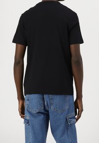 Osoba ubrana w czarną koszulę z krótkim rękawem i niebieskie spodenki dżinsowe cargo, pokazana od tyłu na jednolitym tle.