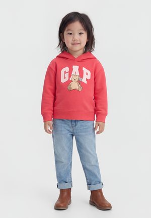 Dziecko stoi ubrane w czerwoną bluzę z kapturem z misiem i logo GAP, jasnoniebieskie podwinięte jeansy i brązowe buty na białym tle.