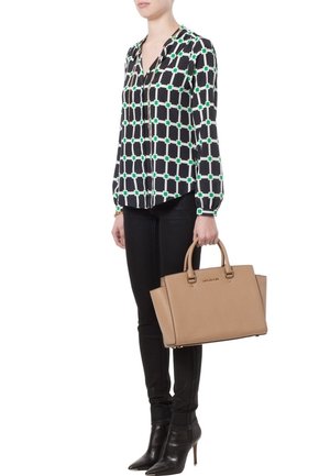 Femme portant un chemisier à motif géométrique noir et blanc, un pantalon noir et des bottines noires, tenant un sac beige Michael Kors.