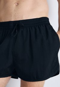 Μαύρες μαγιώ σορτς από ελαφρύ υλικό, με ελαστική μέση και κορδόνι, πλαϊνές τσέπες και λεία υφή.