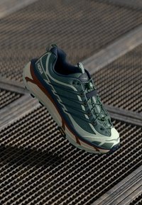 Grüner Sportschuh mit strukturiertem Obermaterial, mehreren Schnürschlaufen, einer gepolsterten Sohle und kontrastierenden braunen Akzenten. Lebendiges und ergonomisches Design.