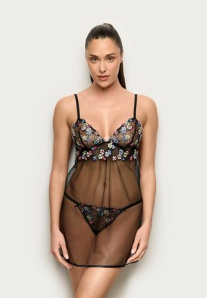 Mujer con el pelo largo, vistiendo un baby doll de lencería negra translúcida y un conjunto a juego de sujetador y bragas con bordado floral, de pie contra un fondo claro y liso.