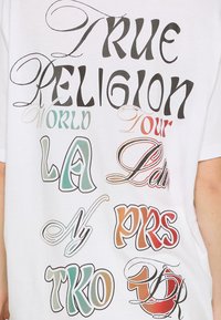 Biała koszulka z różnorodnym, stylizowanym napisem w kolorach czarnym, zielonym, pomarańczowym i brązowym, z wyraźnym napisem "TRUE RELIGION" na górze.