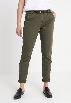 Person wearing rolled-up olive green pants with a braided belt, white shirt, and black slip-on shoes standing against a plain white background.