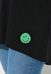 Zwarte stof met een kleine groene glimlachende face patch genaaid nabij de zoom, gecombineerd met lichtblauwe spijkerbroeken.