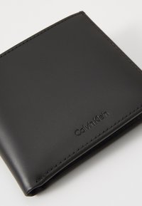 Portafoglio in pelle nera con una texture liscia, caratterizzato da un logo impresso "Calvin Klein" e cuciture ordinate lungo i bordi.