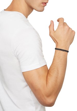 Black woven bracelet featuring a textured design, worn on a bare wrist, contrasts with a white t-shirt in a side-profile view.