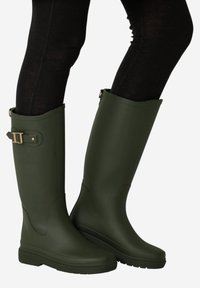 Botas de goma verdes oscuras hasta la rodilla con hebilla lateral, usadas sobre leggings negros sobre un fondo blanco.