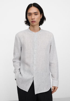 Jeune personne aux cheveux foncés mi-longs, portant une chemise boutonnée gris clair à manches longues et un pantalon foncé sur fond blanc.