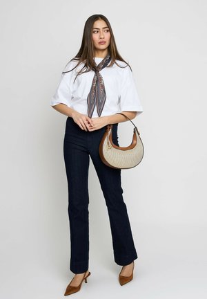 Frau trägt ein weißes kurzärmeliges Oberteil, marineblaue Hose, braune Schuhe mit Absatz, ein gemustertes Halstuch und hält eine beige-braune Handtasche.