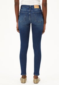 Jean skinny en denim bleu foncé avec une texture lisse, doté d'une taille haute et de poches arrière, fini avec une étiquette en cuir portant la marque.