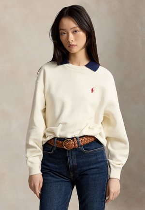 Jeune femme aux longs cheveux foncés portant un pull écru avec un col bleu marine, un jean bleu et une ceinture tressée marron, debout immobile.
