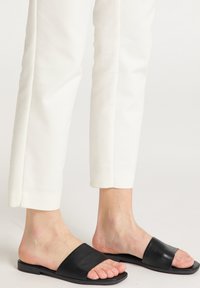 Zwarte leren slip-ons met een vlakke zool, voorzien van een brede, gladde band en een minimalistisch design. Gemaakt in combinatie met witte taps toelopende broeken.