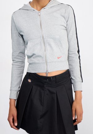 Femme portant un sweat à capuche court gris clair zippé avec des bandes noires sur les manches et une jupe plissée noire avec un patch logo à la taille.