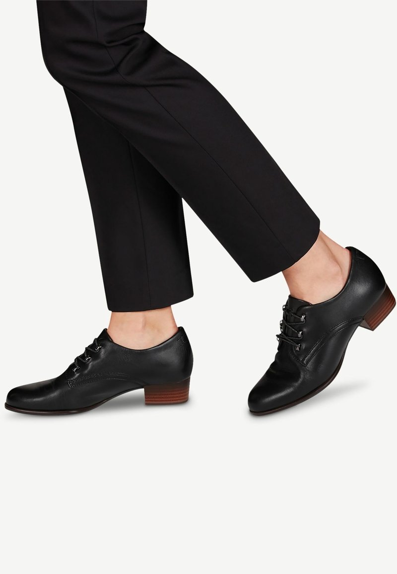 Scarpe stringate in pelle nera con tacco basso, caratterizzate da una superficie liscia e punta arrotondata. Accenti includono cuciture decorative e occhielli in metallo.