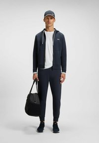 Mørkeblå atletisk outfit med en lynlåsjakke, tætsiddende bukser og en hvid t-shirt. Sort sportstaske, blå sneakers og en matchende kasket.