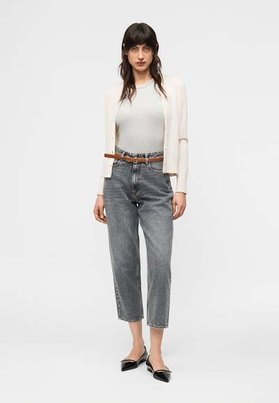 Γυναίκα που φοράει ανοιχτό μπεζ ζακέτα πάνω από γκρι τοπ, γκρι τζιν με ψηλή μέση και καφέ ζώνη, καθώς και μαύρες μυτερές μπαλαρίνες, στέκεται μπροστά σε λευκό φόντο.