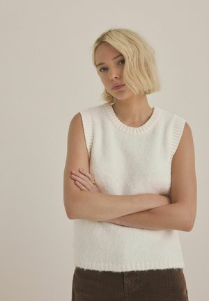 White sleeveless sweater with a ribbed neckline, textured fabric, and a straight hem, paired with brown corduroy pants.