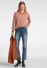 Top de manga larga rojo y blanco con patrón, vaqueros azules, botas negras de tobillo, sosteniendo una chaqueta de cuero marrón. Diseño simple con un corte relajado.