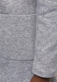 Grått stickat tyg med en mjuk textur, som har en ficka i lappstil och en design med stackade ärmar. Materialet verkar sträckt och bekvämt.