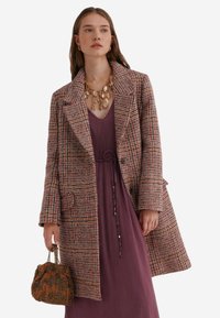 Kvinde iført en ternet frakke over en lilla kjole, der holder en brun perlehandtaske og er accessoriseret med et stort halskæde med guldbelægning.