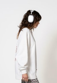 Felpa bianca oversize con maniche a costine, indossata con pantaloni grigi e neri a fantasia, e cuffie argentate appoggiate sui capelli ricci.