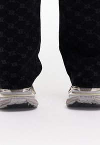 Pantalon noir à motif avec un design de logo répété, associé à des baskets gris et blanc avec des semelles texturées et des accents réfléchissants.