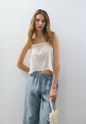 Top corto blanco con bordado de encaje, combinado con pantalones de lino azul claro con cordón. La modelo sostiene un pequeño bolso texturizado crema.
