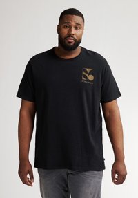 Camiseta de algodón negra con un logo gráfico dorado en el área del pecho izquierdo, cuello redondo y mangas cortas. Lleva puestas jeans grises.