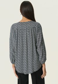 Svart och vit prickig blus med en rynkad baksida, rund hals och långa ärmar, tillverkad av lättviktsmaterial.