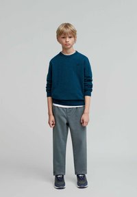 Sudadera verde azulado de manga larga y cuello redondo, superpuesta sobre una camisa blanca; combinada con pantalones grises y zapatillas azul marino.