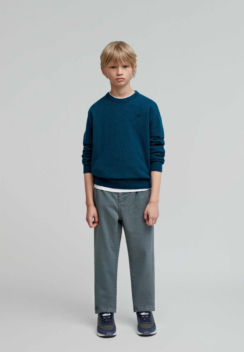 Sudadera verde azulado de manga larga y cuello redondo, superpuesta sobre una camisa blanca; combinada con pantalones grises y zapatillas azul marino.