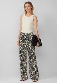 Witte geribbelde tanktop, zwart en crèmekleurige broek met een bladerenpatroon en een zwarte gestructureerde handtas. Platte sandalen maken de look af.