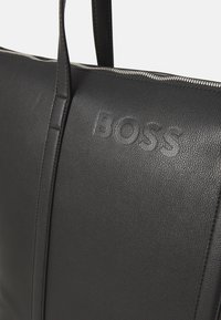 Svart skinn toteveske med en myk tekstur, preget logo og detaljer i sølvglidelås langs toppen. Glatt, strukturert design.