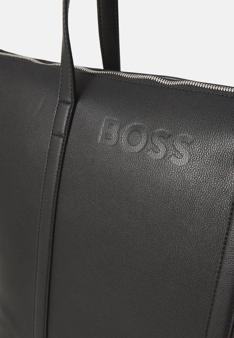 Sac fourre-tout en cuir noir avec une texture douce, logo en relief et détails de fermeture à glissière argentés sur le dessus. Design lisse et structuré.