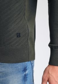 Donkergroene textuurtrui, met diagonale breipatronen en een geribbelde onderrand. Klein logo-embleem aan de zijkant, gecombineerd met lichtblauwe jeans.