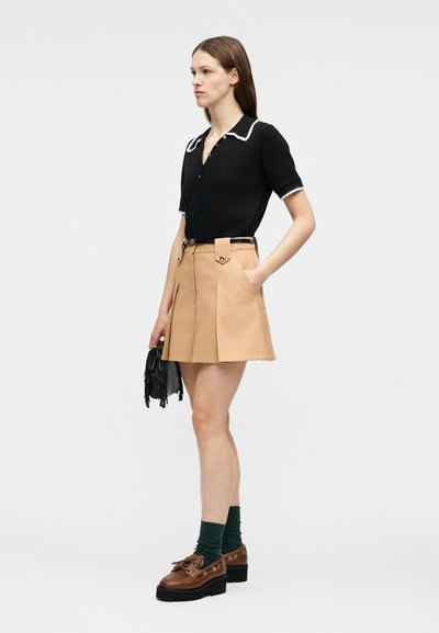 Γυναίκα στέκεται φορώντας μπεζ φούστα ψηλόμεση με ζώνη, μαύρο μπλουζάκι με κοντά μανίκια και λευκή επένδυση, πράσινες κάλτσες, καφέ παπούτσια με τακούνι πλατφόρμα, κρατώντας μαύρη τσάντα με κρόσσια.