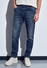 Hombre vestido con jeans de mezclilla azul oscuro y zapatillas blancas, con una mano en el bolsillo, de pie contra una pared blanca y un suelo gris.