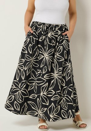 Frau trägt einen schwarzen Maxirock mit großem cremefarbenem Blumenmuster, elastischem Bund und vorderen Taschen, kombiniert mit weißen Sandalen und einem weißen Oberteil.