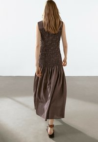 Lång, ärmlös klänning med en volangad bodice i mörkbrunt, flödande kjol av texturerad tyg, buren med svarta klackar med remmar.