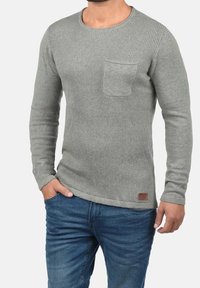Grauer, gerippter Pullover mit langen Ärmeln und Rundhalsausschnitt, mit einer Brusttasche und einem kontrastierenden braunen Lederetikett am Saum.